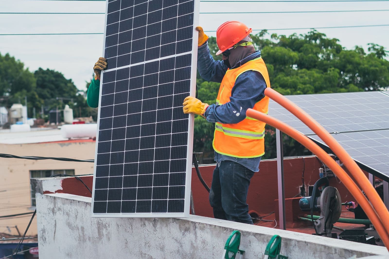 A technician in safety gear carrying a solar panel across a rooftop.