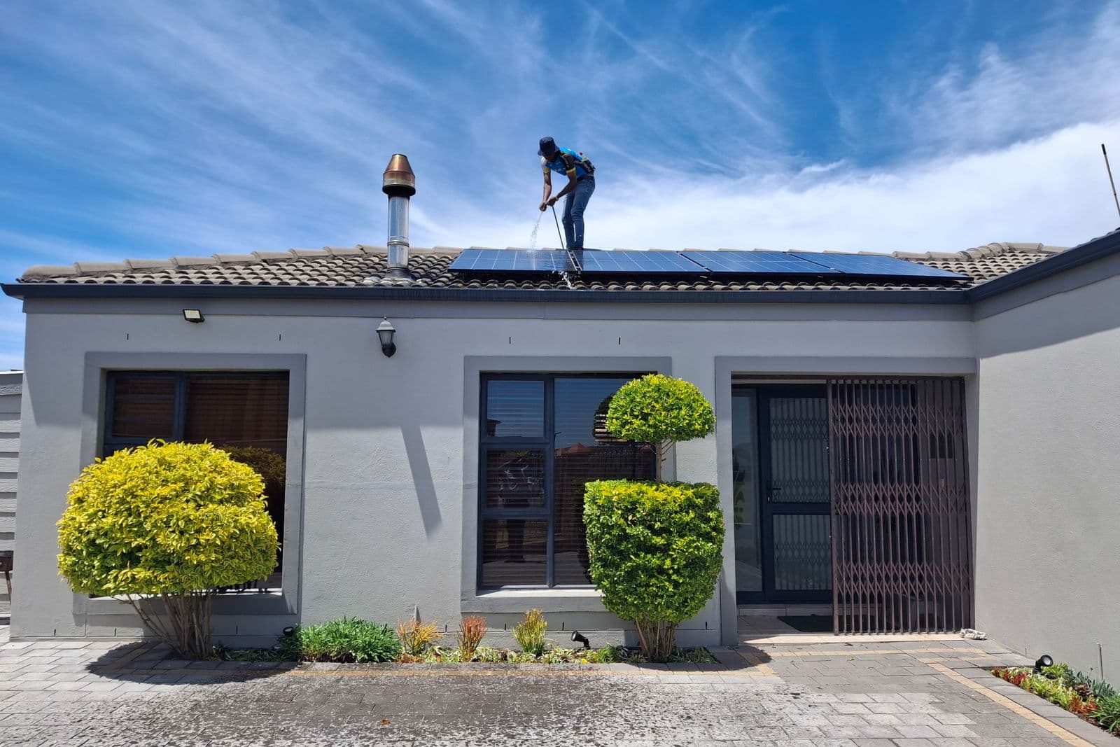 A single storey house with a solar array on its tiled roof, and a worker on the roof rinsing the panels.