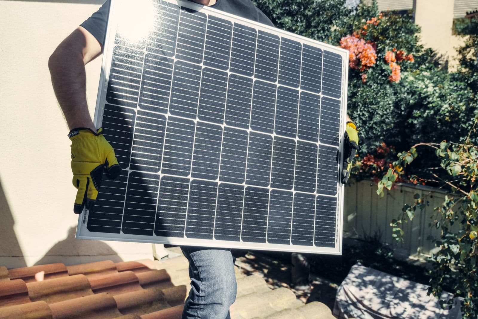 An installer carrying a solar panel across a tiled roof, the panel filling most of the frame.