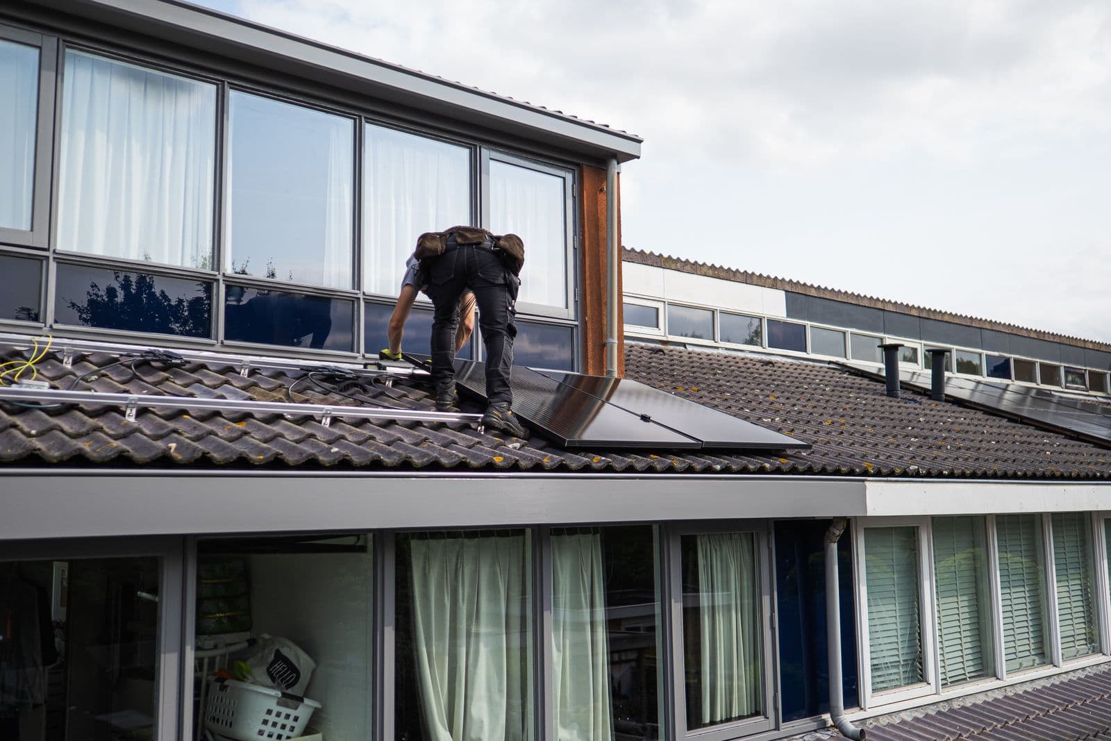 An installer on a tiled residential roof fitting a black solar panel onto mounting rails.