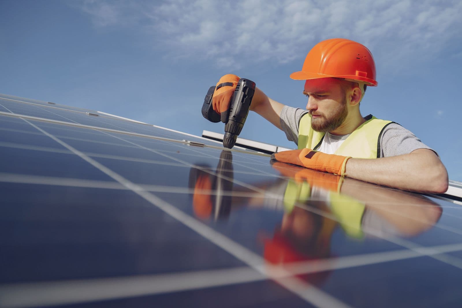 An electrician in a hard hat fixing a solar panel with a drill.